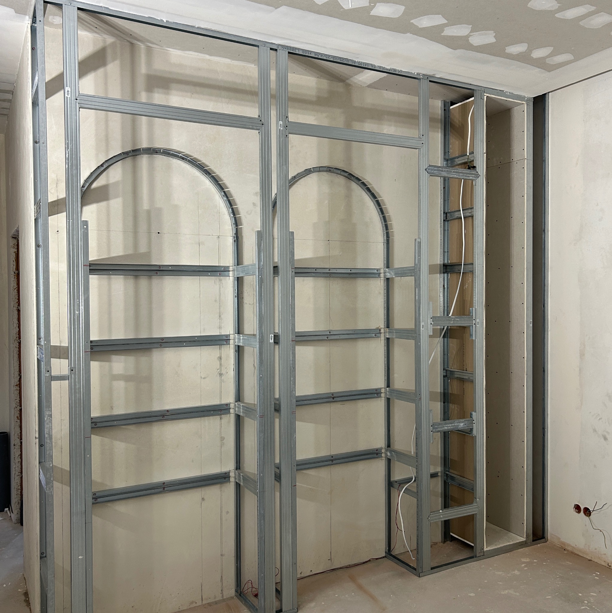 Konstrukcja metalowa z profili do zabudowy ścianki działowej z płyt kartonowo-gipsowych, widoczne profile pionowe i poziome tworzące szkielet z wyciętymi łukami, częściowo obłożona płytami...