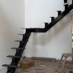 Kul-Spaw - Czarne metalowe schody bez balustrady w trakcie montażu w surowym wnętrzu, osoba wchodzi po schodach, widoczne kartony i resztki materiałów budowlanych na podłodze.