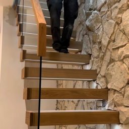 Kul-Spaw - Nowoczesne schody drewniane z podświetleniem LED i szklaną balustradą, zamontowane w Rawiczu, przylegające do ściany z kamienia dekoracyjnego, osoba wchodzi po schodach.