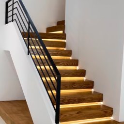 Kul-Spaw - Nowoczesne drewniane schody z podświetleniem LED pod stopniami i czarną, minimalistyczną balustradą, w jasnym wnętrzu.