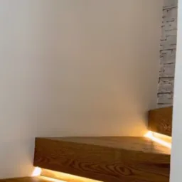 Nowoczesne schody z drewna z podświetleniem LED, widok z dołu, jasne drewno z wyraźnym usłojeniem, biała ściana w tle.