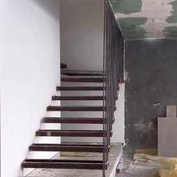 Kul-Spaw - Wnętrze w trakcie wykańczania: konstrukcja schodów z metalowymi stopniami i minimalistyczną, pionową balustradą z prętów, widoczne niedokończone ściany i podłoga.