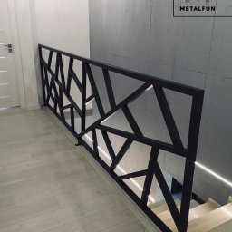 Kul-Spaw - Nowoczesna, czarna balustrada schodowa z geometrycznym wzorem, oświetlona listwą LED, na tle betonowej ściany i jasnej podłogi. Widoczny fragment schodów.