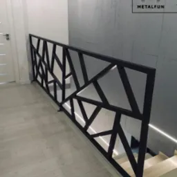 Nowoczesna, czarna balustrada schodowa z geometrycznym wzorem, oświetlona listwą LED, na tle betonowej ściany i jasnej podłogi. Widoczny fragment schodów.