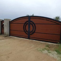 Kul-Spaw - Brama wjazdowa z brązowymi panelami i czarnymi, okrągłymi zdobieniami, osadzona na betonowych słupkach. Widok na wjazd i fragment zieleni.