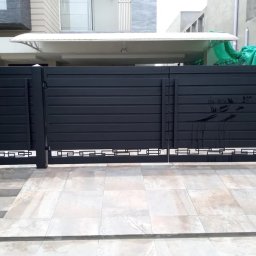 Kul-Spaw - Nowoczesna, pozioma brama wjazdowa w kolorze czarnym, z dekoracyjnym, ażurowym wykończeniem u dołu. Widoczne schody i fragment domu w tle.