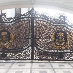 Kuta, ozdobna brama wjazdowa z herbem w złotym wieńcu, widziana od frontu. Bogate zdobienia roślinne, symetryczny układ elementów, jasne tło budynku.