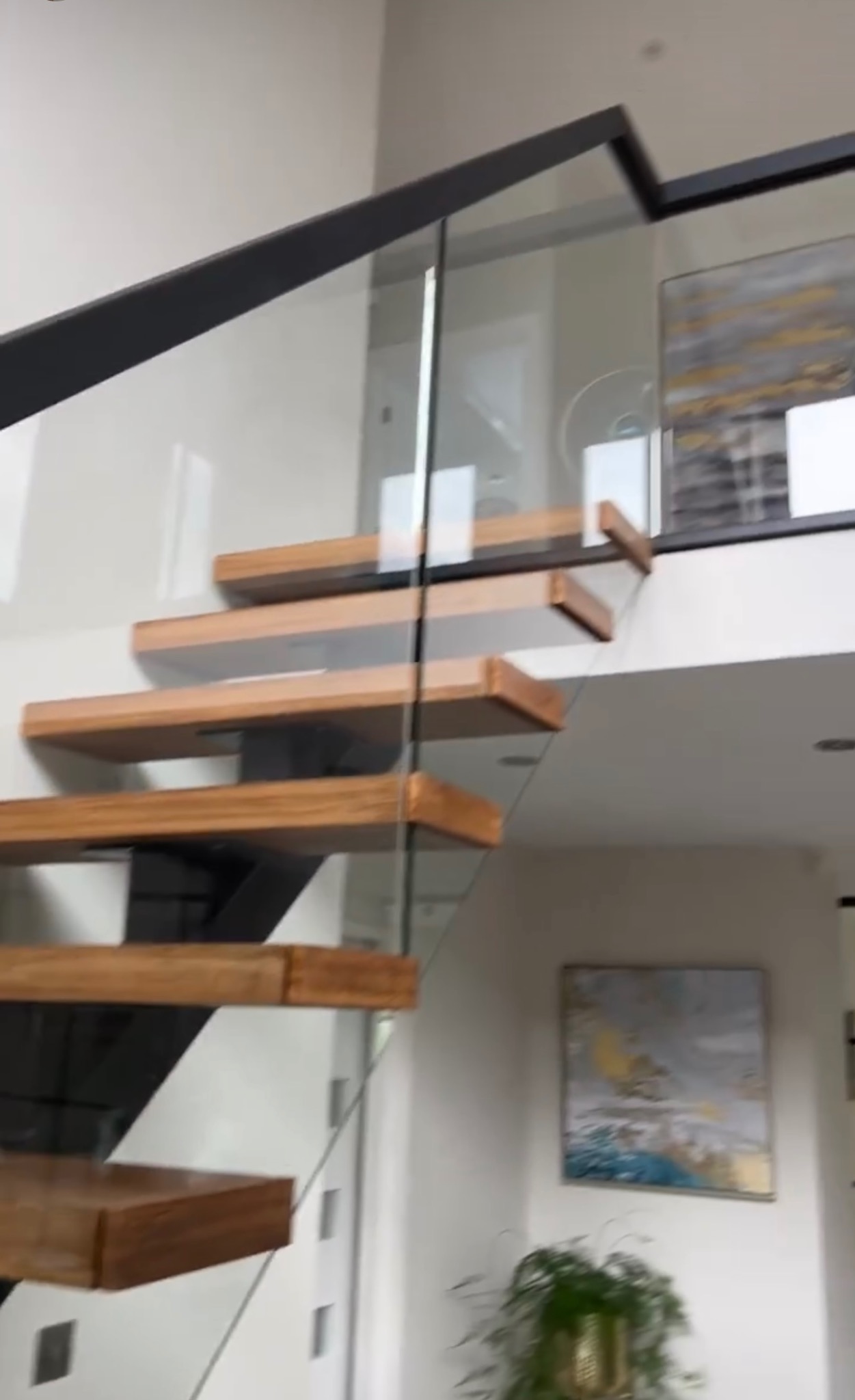 Nowoczesne schody z drewnianymi stopniami i szklaną balustradą, wspornikowe stopnie przymocowane do ściany, czarna metalowa konstrukcja, jasne wnętrze.