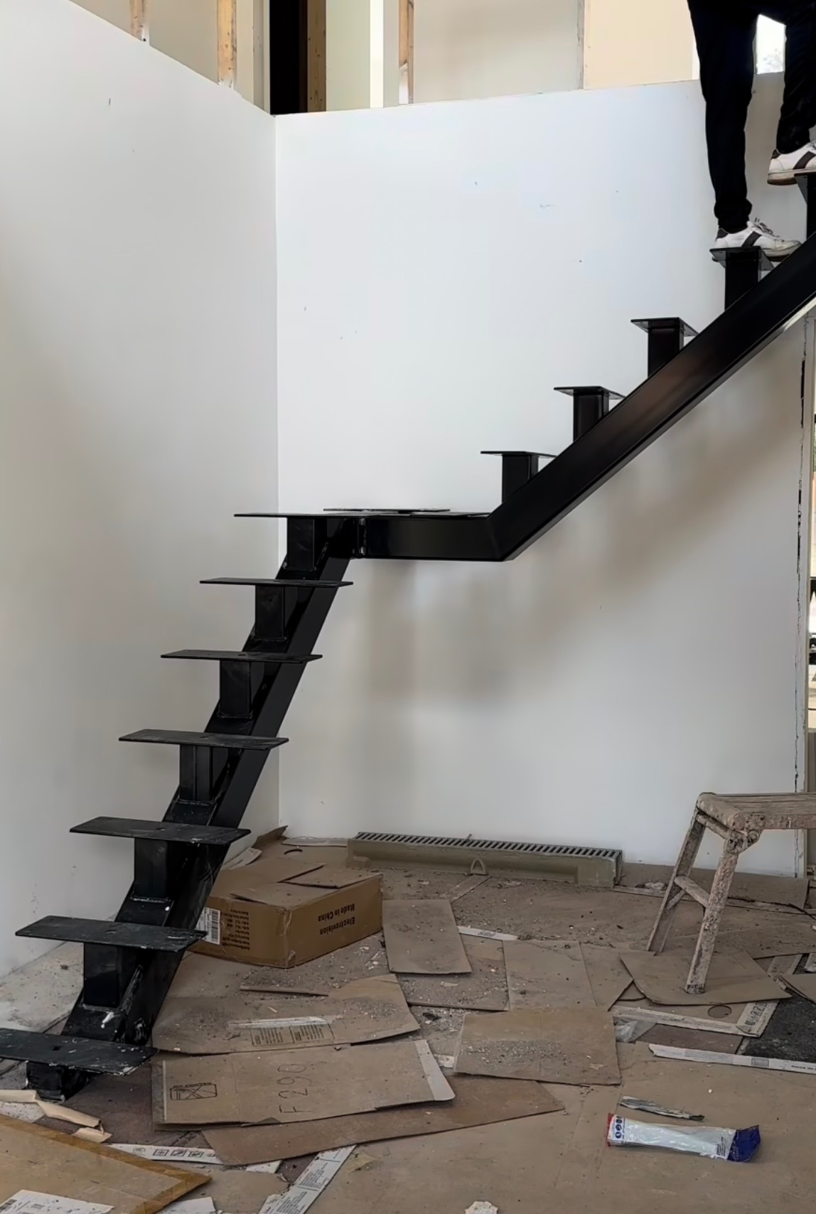 Czarne metalowe schody bez balustrady w trakcie montażu w surowym wnętrzu, osoba wchodzi po schodach, widoczne kartony i resztki materiałów budowlanych na podłodze.