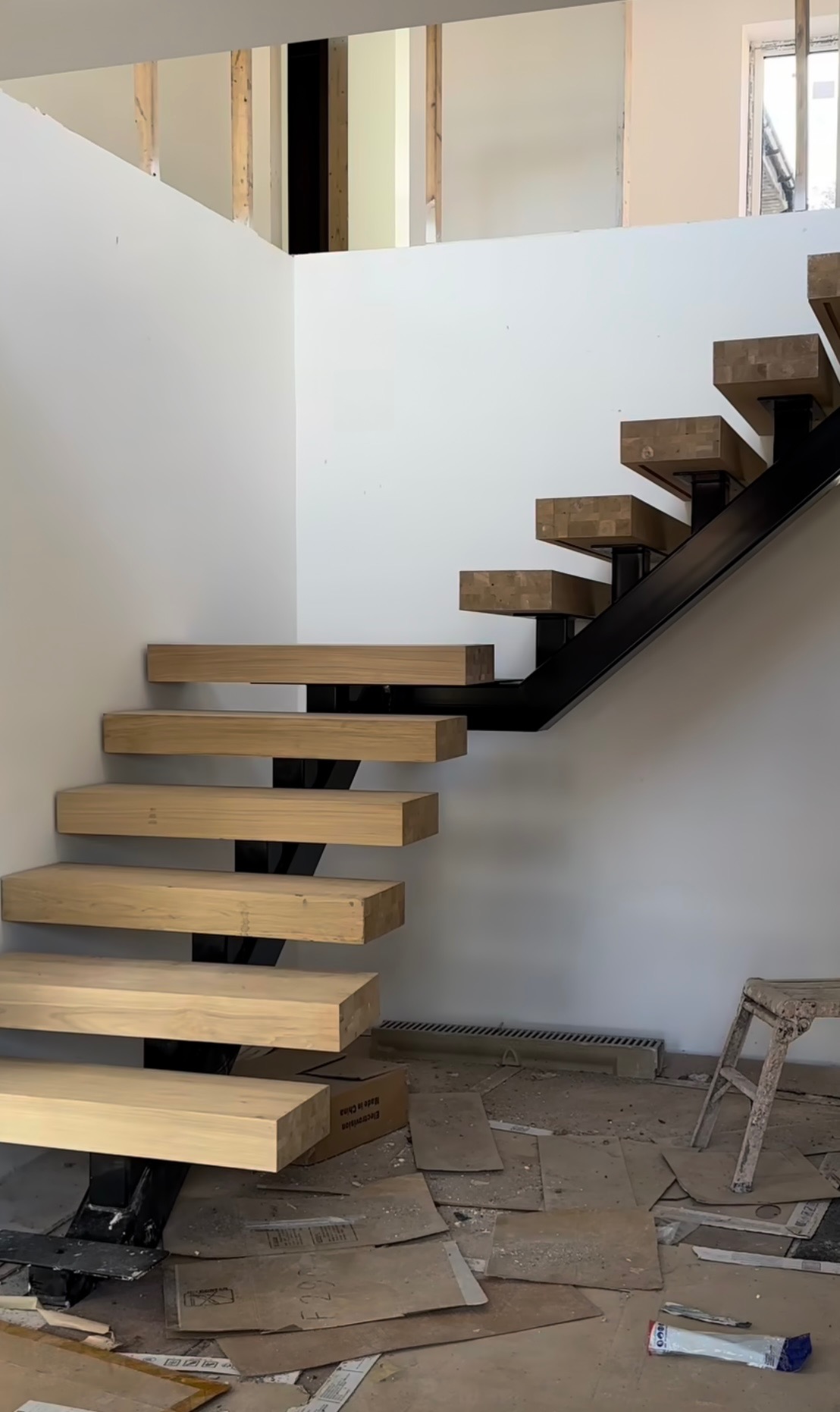 Surowe drewniane stopnie schodów wsparte na czarnej metalowej konstrukcji, w trakcie montażu, widoczne elementy wykończeniowe podłogi i ściany.