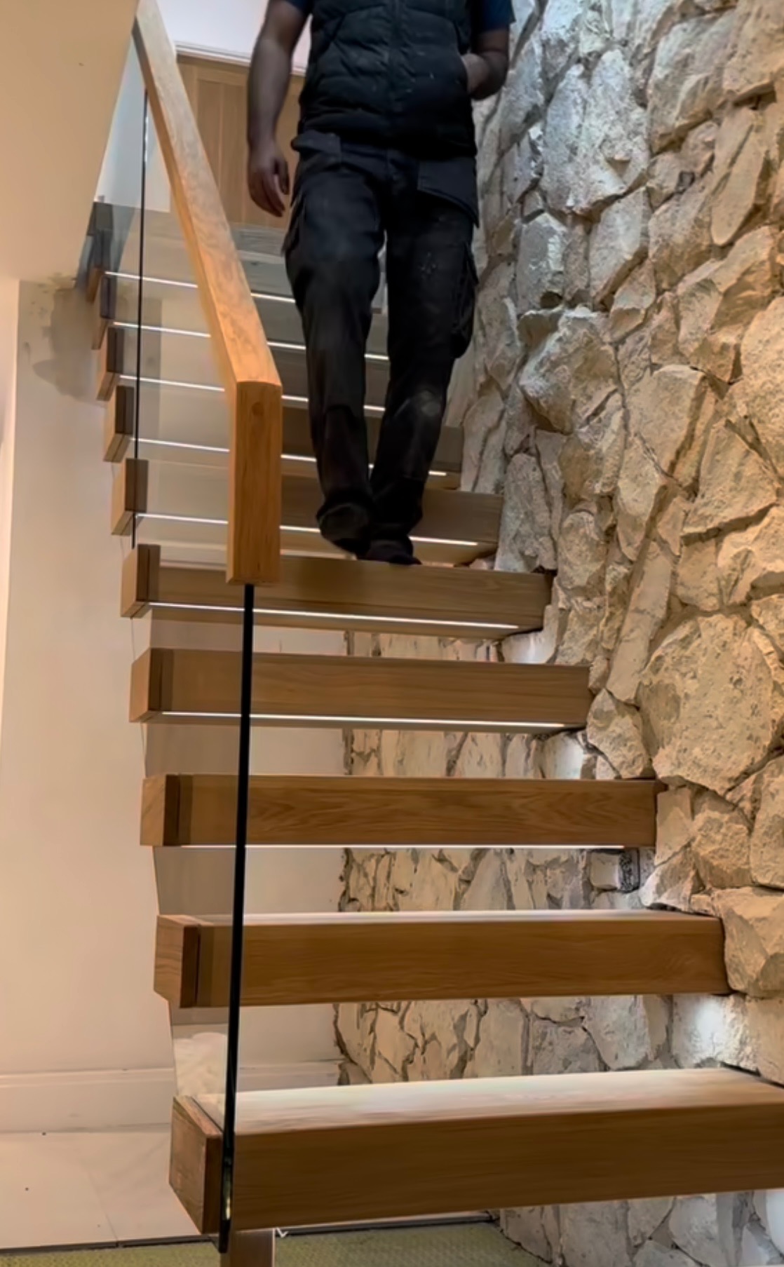 Nowoczesne schody drewniane z podświetleniem LED i szklaną balustradą, zamontowane w Rawiczu, przylegające do ściany z kamienia dekoracyjnego, osoba wchodzi po schodach.