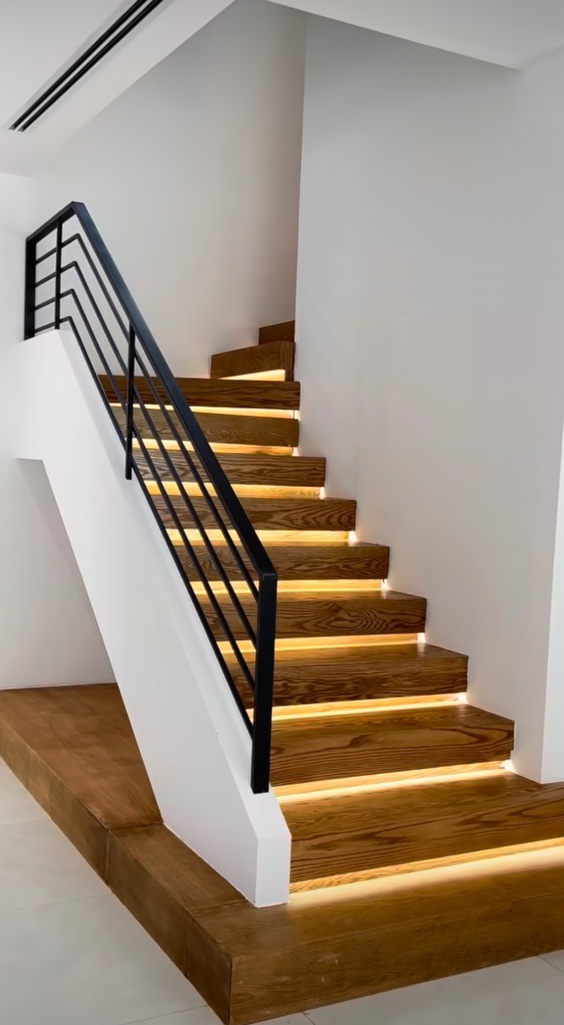 Nowoczesne drewniane schody z podświetleniem LED pod stopniami i czarną, minimalistyczną balustradą, w jasnym wnętrzu.