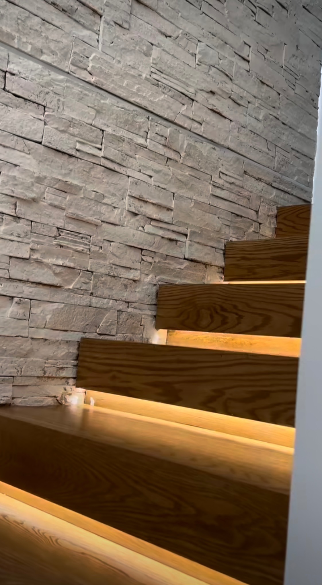 Drewniane schody z podświetleniem LED, przylegające do ściany wyłożonej dekoracyjnym kamieniem w odcieniach bieli i szarości.
