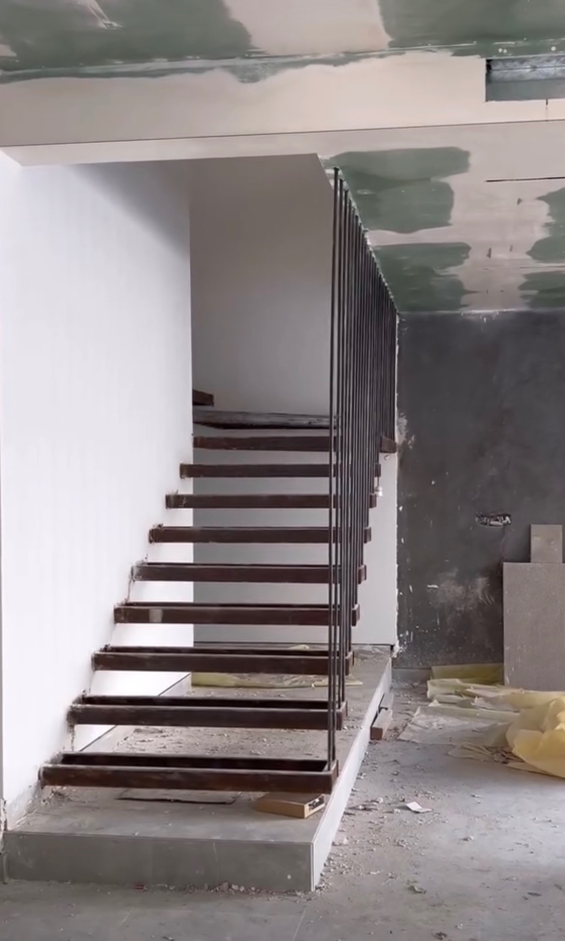 Wnętrze w trakcie wykańczania: konstrukcja schodów z metalowymi stopniami i minimalistyczną, pionową balustradą z prętów, widoczne niedokończone ściany i podłoga.