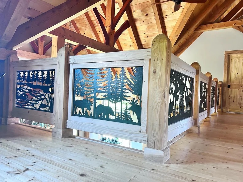 Drewniana balustrada z metalowymi panelami dekoracyjnymi przedstawiającymi motywy leśne: zwierzęta (łoś, niedźwiedź) i drzewa. Ustawiona na drewnianym poddaszu.