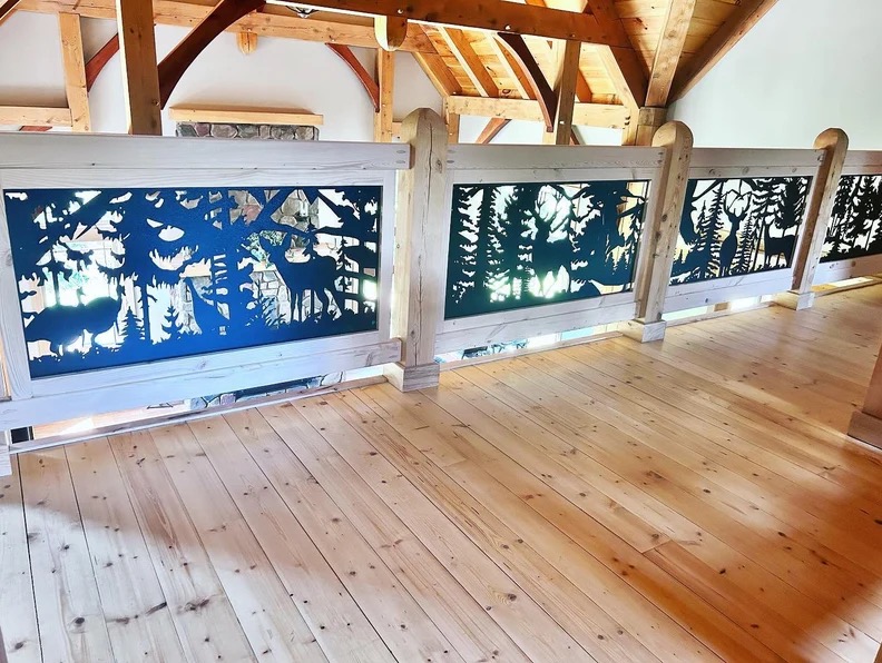 Drewniana balustrada z metalowymi panelami wycinanymi laserowo, przedstawiającymi motywy leśne: jelenie, drzewa i wilki, oświetlona naturalnym światłem w rustykalnym wnętrzu.