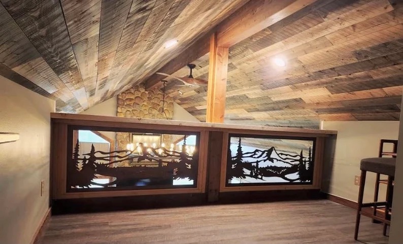 Drewniana balustrada z metalowymi panelami o wzorach górskich i leśnych, widoczna na tle drewnianego sufitu i kamiennej ściany. Unikalny detal w loftowym wnętrzu.