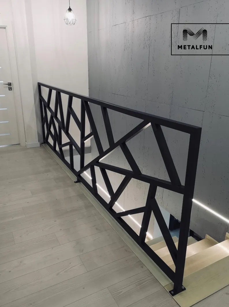 Nowoczesna, czarna balustrada schodowa z geometrycznym wzorem, oświetlona listwą LED, na tle betonowej ściany i jasnej podłogi. Widoczny fragment schodów.