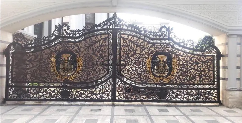 Kuta, ozdobna brama wjazdowa z herbem w złotym wieńcu, widziana od frontu. Bogate zdobienia roślinne, symetryczny układ elementów, jasne tło budynku.