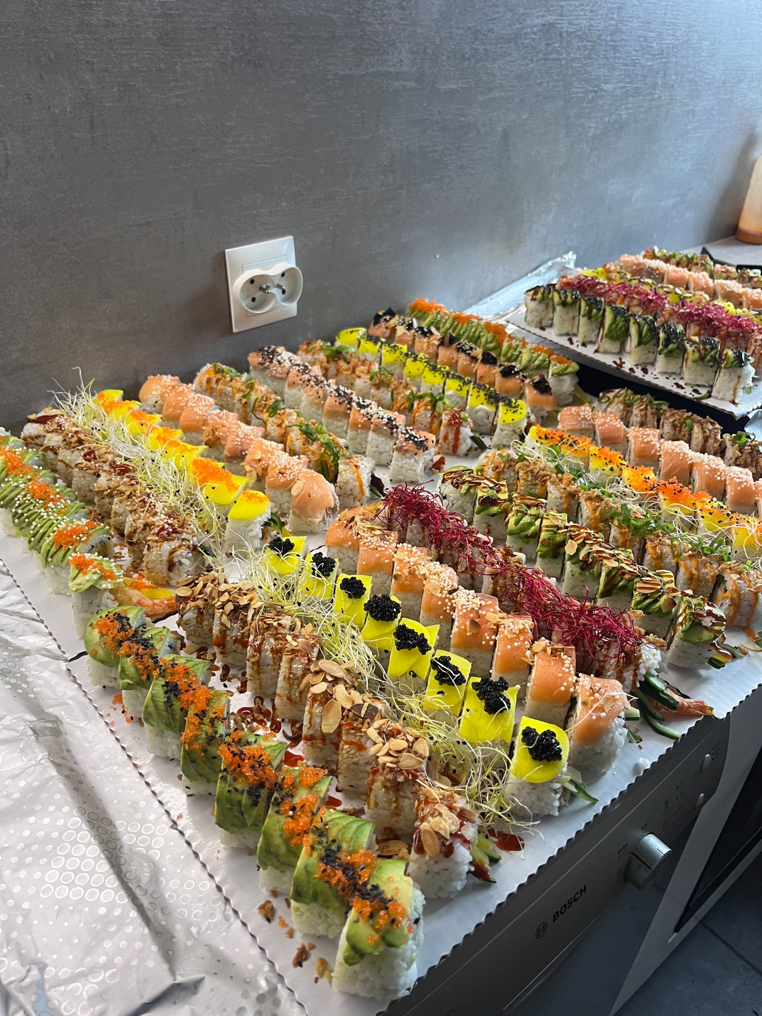 Bogaty wybór różnorodnych rolek sushi, ułożonych rzędami na stole z folią dekoracyjną, widoczne różne dodatki: kawior, łosoś, awokado, posypki, kiełki.