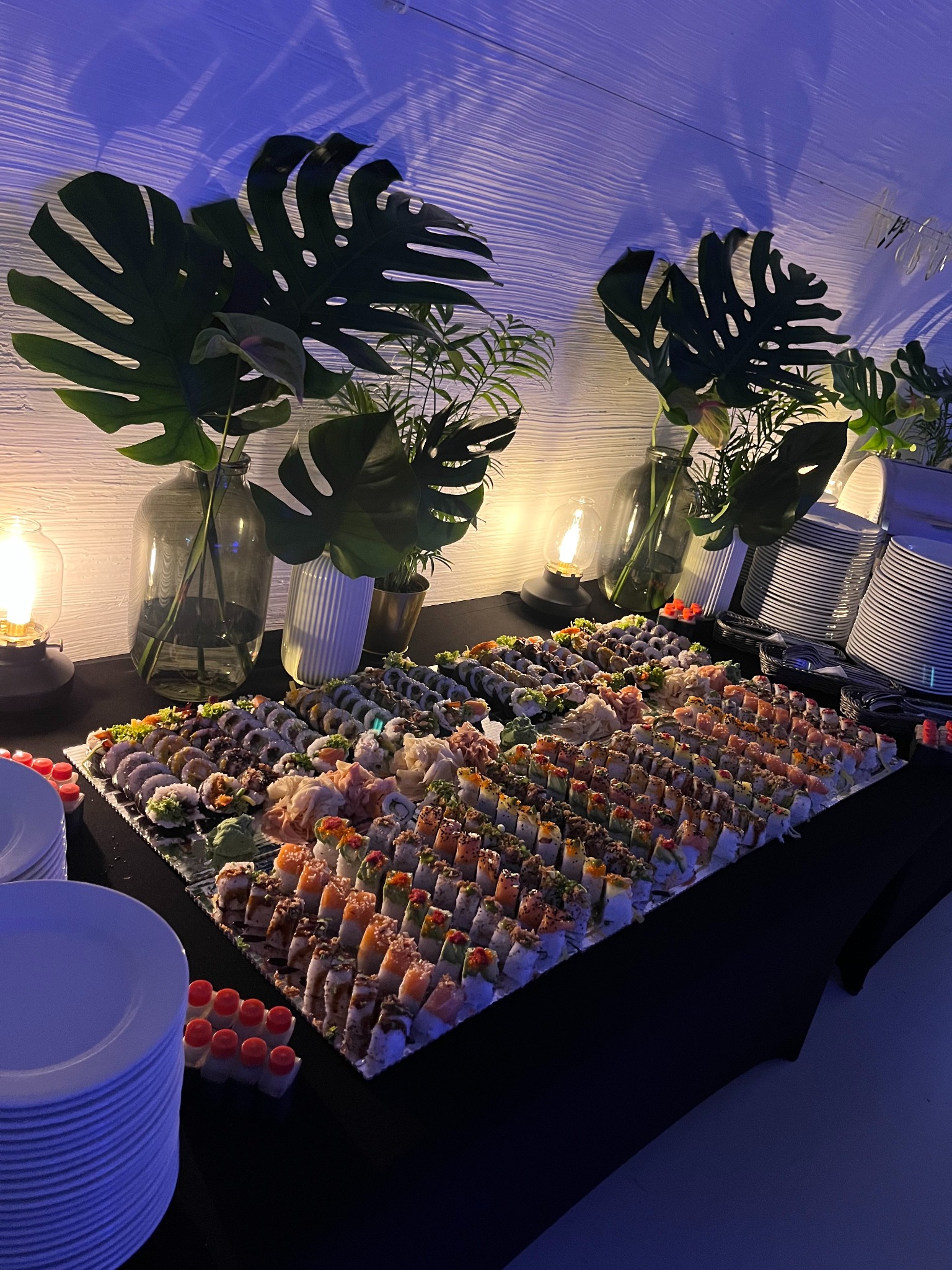 Stół cateringowy z różnorodnym sushi, dekoracjami roślinnymi w szklanych wazonach i stosami białych talerzy, oświetlony nastrojowym, niebieskawym światłem.