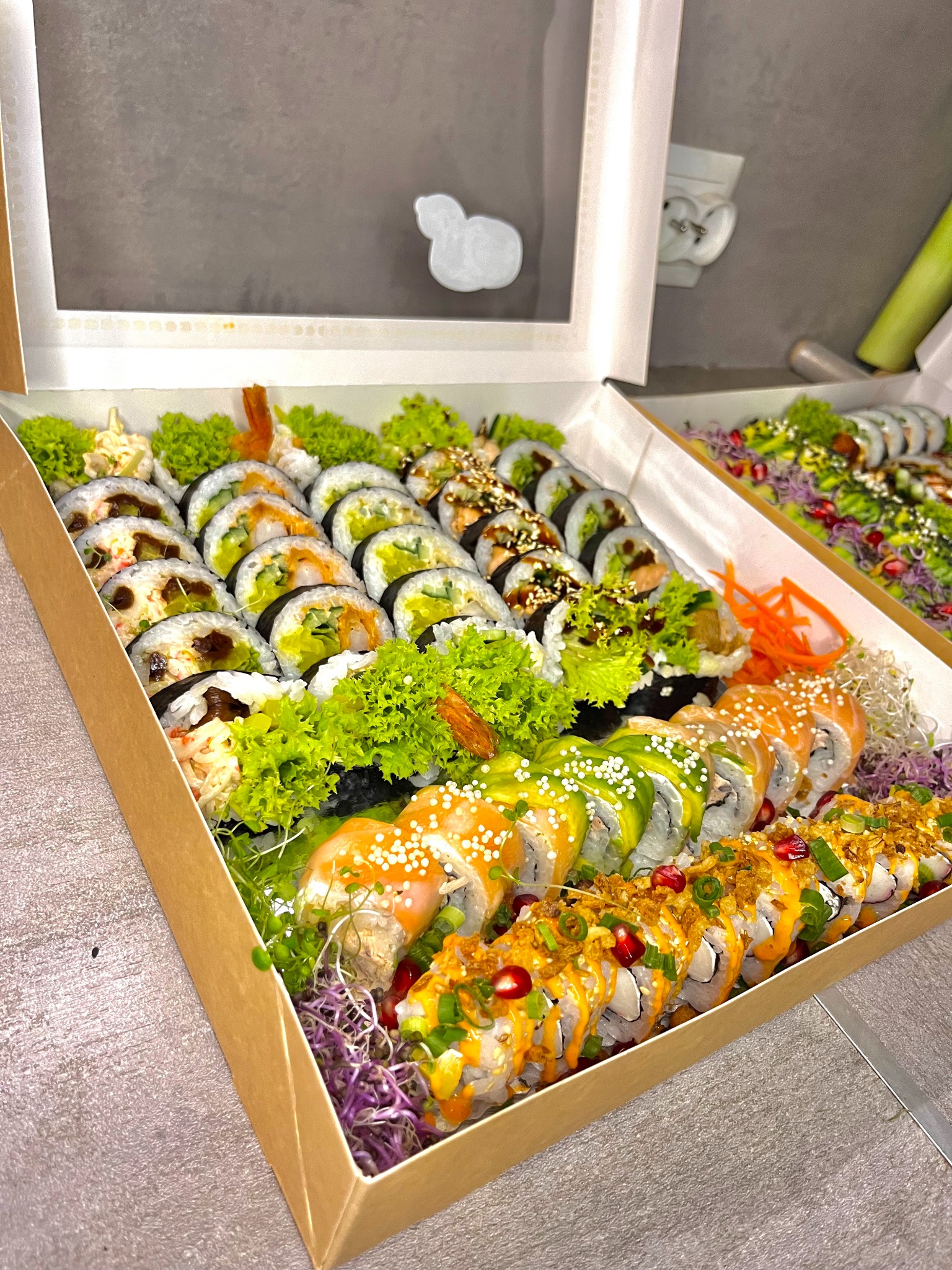 Zestaw różnorodnych rolek sushi w tekturowym pudełku z przezroczystym wieczkiem, udekorowany sałatą, kiełkami i marchewką, widoczne różne rodzaje nadzień i posypek.
