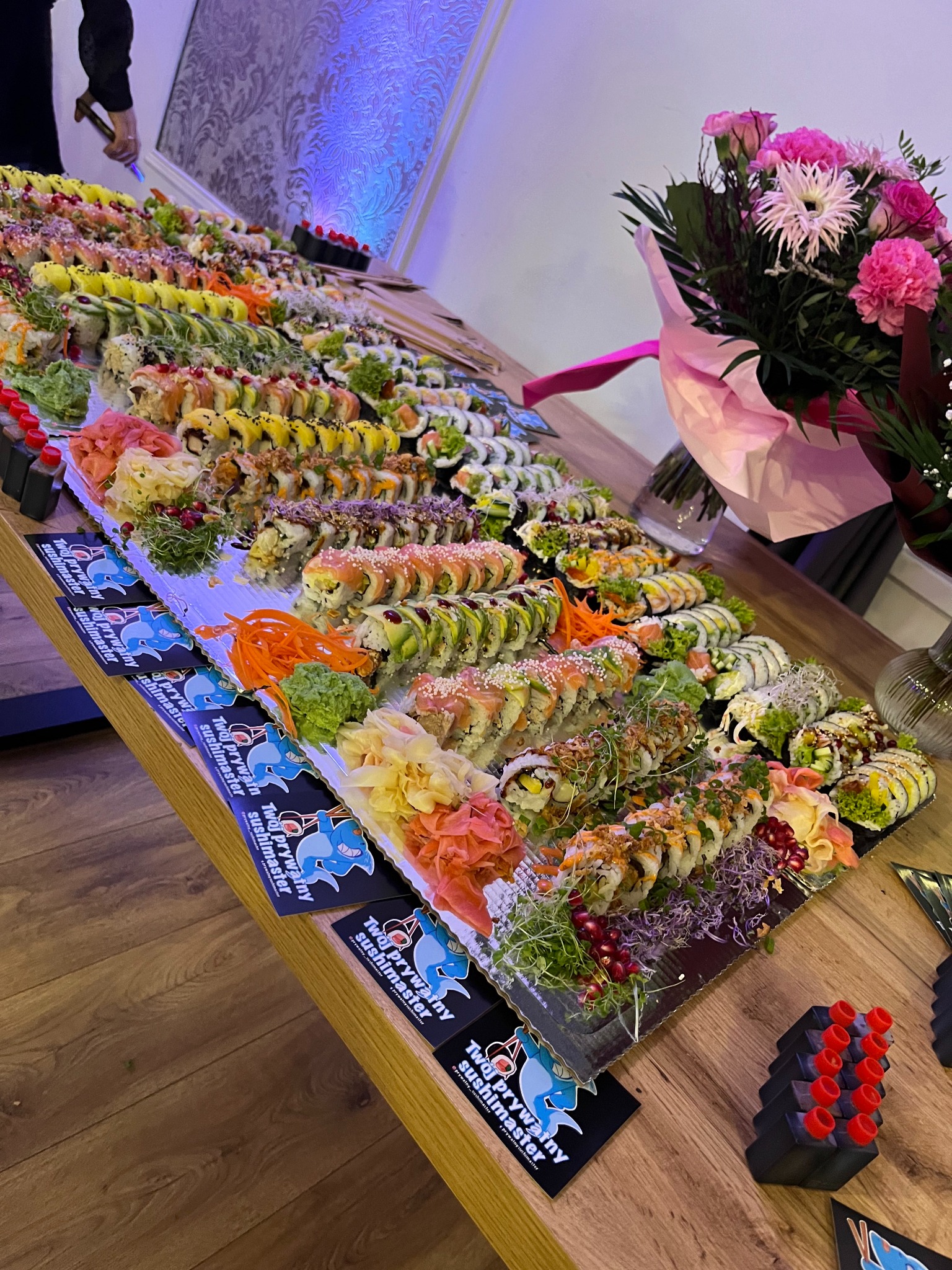 Stół z bogatym zestawem sushi, różnorodne rolki, imbir, wasabi, sos sojowy, dekoracja kwiatowa w tle, wizytówki firmy cateringowej na stole.
