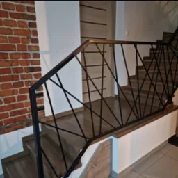 Drewniane schody z czarną, geometryczną balustradą z metalowych prętów, widoczne na tle ceglanej ściany i jasnych drzwi wewnętrznych.