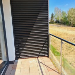 DiM Studio - Nowoczesny balkon w Łukawcu z czarną, poziomą przesłoną przeciwsłoneczną i balustradą ze stali nierdzewnej z wypełnieniem szklanym, widok na plac budowy i drzewa w oddali.
