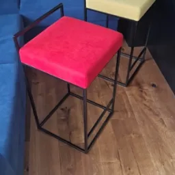 Dwa nowoczesne taborety barowe: jeden z czerwonym siedziskiem, drugi z żółtym, oba z czarną metalową ramą, stoją na drewnianej podłodze obok niebieskiej sofy.