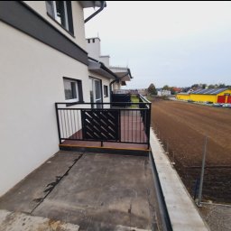 DiM Studio - Czarna balustrada balkonowa z dekoracyjnym wypełnieniem w postaci ukośnych prętów, widok z boku na balkon, w tle pole i budynek gospodarczy.