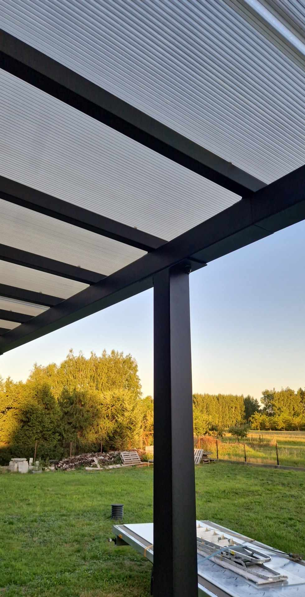 Czarna metalowa pergola z przezroczystym zadaszeniem z widokiem na zielony ogród i błękitne niebo o zmierzchu.