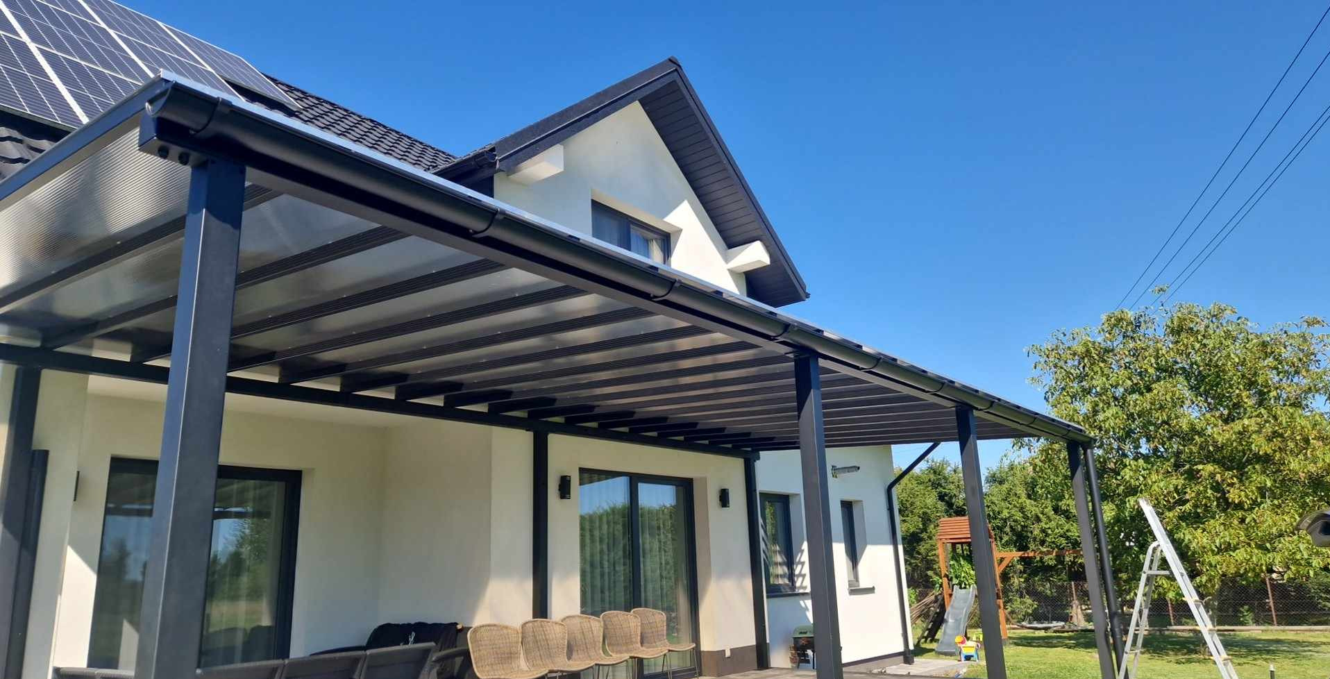 Nowoczesna pergola metalowa z przezroczystym zadaszeniem, przylegająca do białego domu z panelami słonecznymi na dachu, widok z boku na tle błękitnego nieba i zieleni ogrodu.
