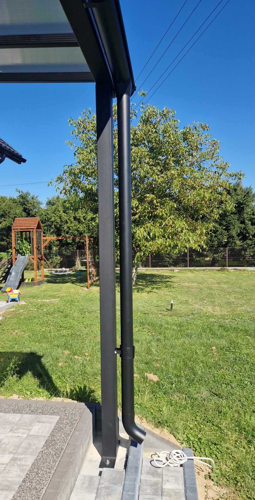 Czarna metalowa konstrukcja z rynną na tle zielonego ogrodu z placem zabaw dla dzieci w słoneczny dzień.