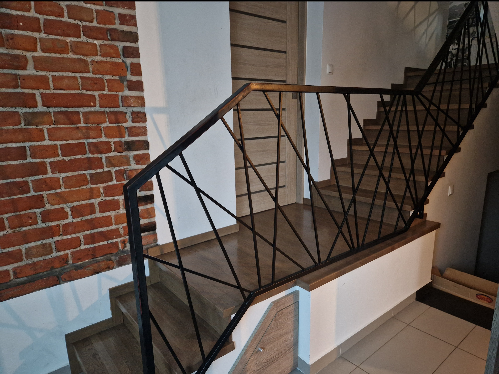 Drewniane schody z czarną, geometryczną balustradą z metalowych prętów, widoczne na tle ceglanej ściany i jasnych drzwi wewnętrznych.
