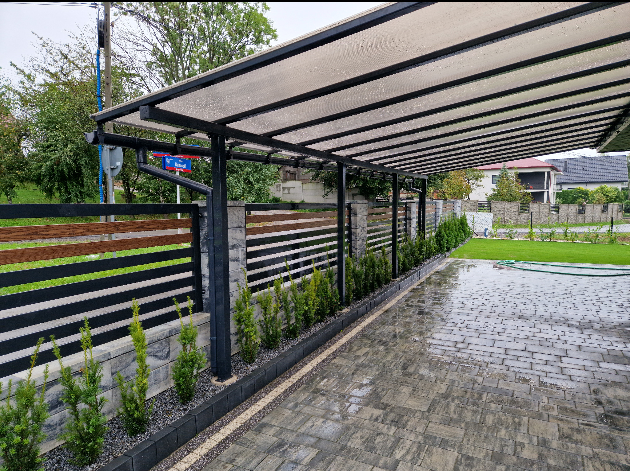 Nowoczesna, czarna pergola metalowa z przezroczystym zadaszeniem nad wybrukowanym podjazdem, obok drewniano-metalowego ogrodzenia i rzędu niskich iglaków.