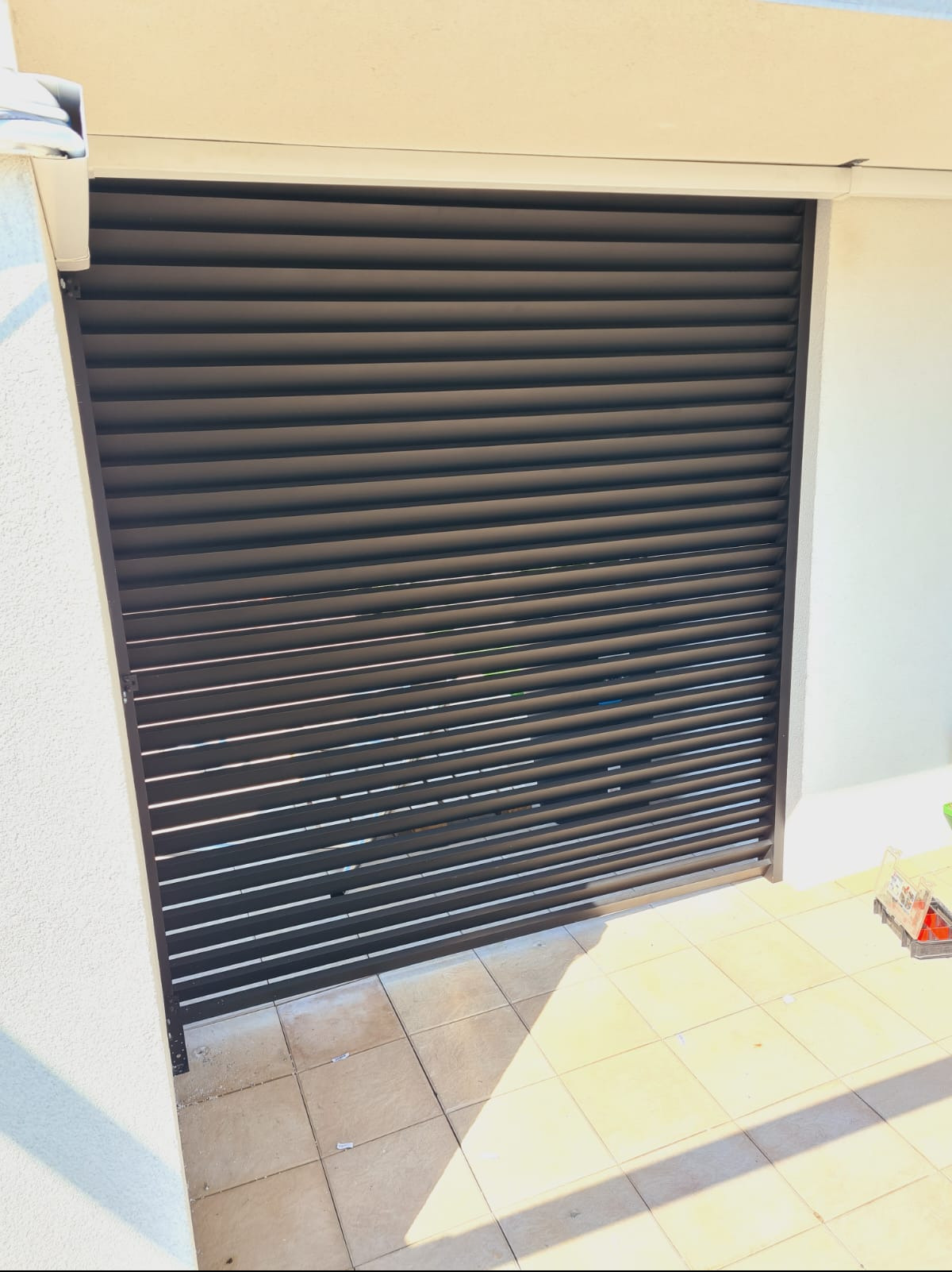 Zewnętrzna, pozioma żaluzja metalowa w kolorze grafitowym, zamontowana na balkonie z jasnymi płytkami, częściowo zasłonięta.