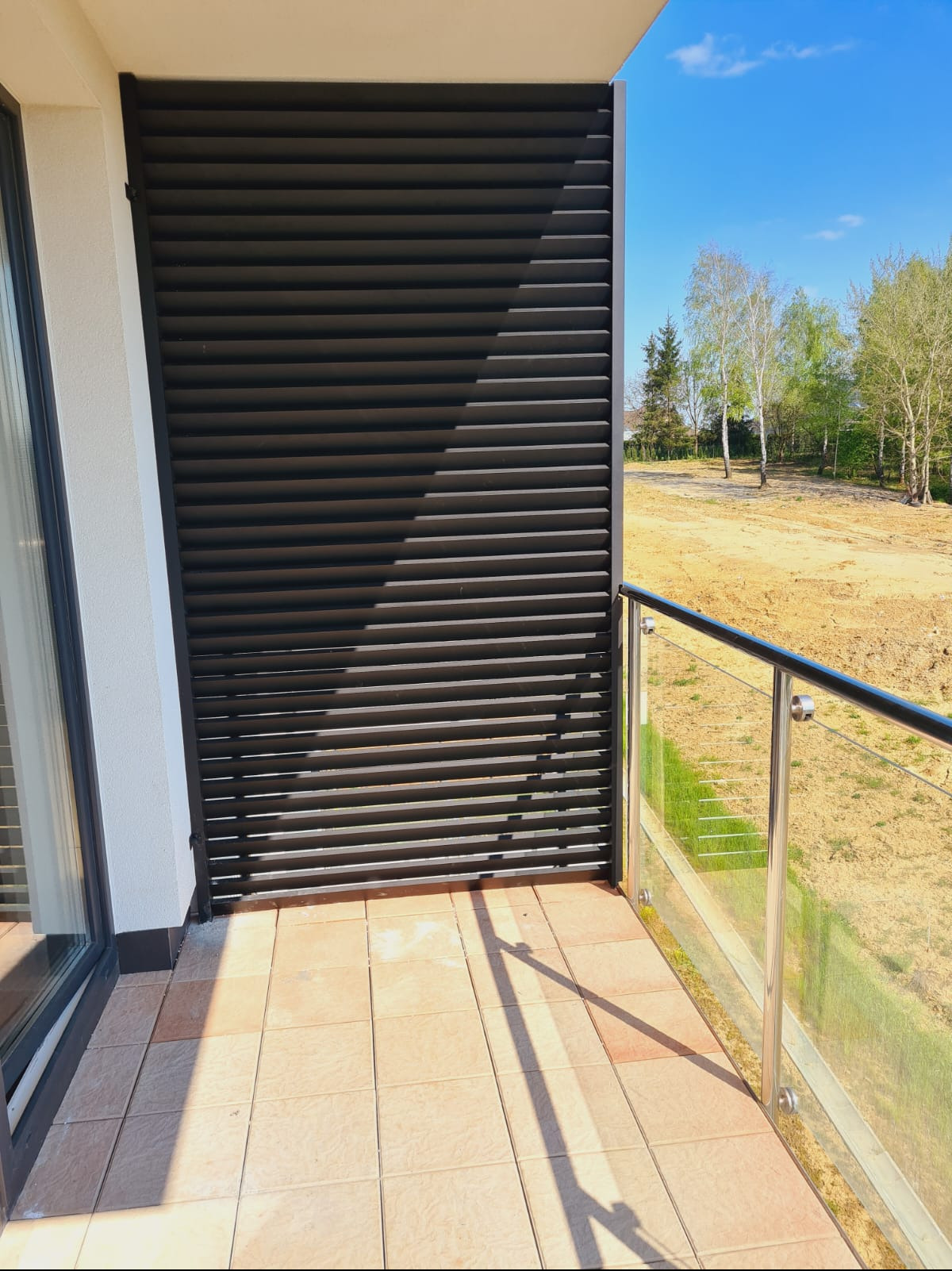 Nowoczesny balkon w Łukawcu z czarną, poziomą przesłoną przeciwsłoneczną i balustradą ze stali nierdzewnej z wypełnieniem szklanym, widok na plac budowy i drzewa w oddali.