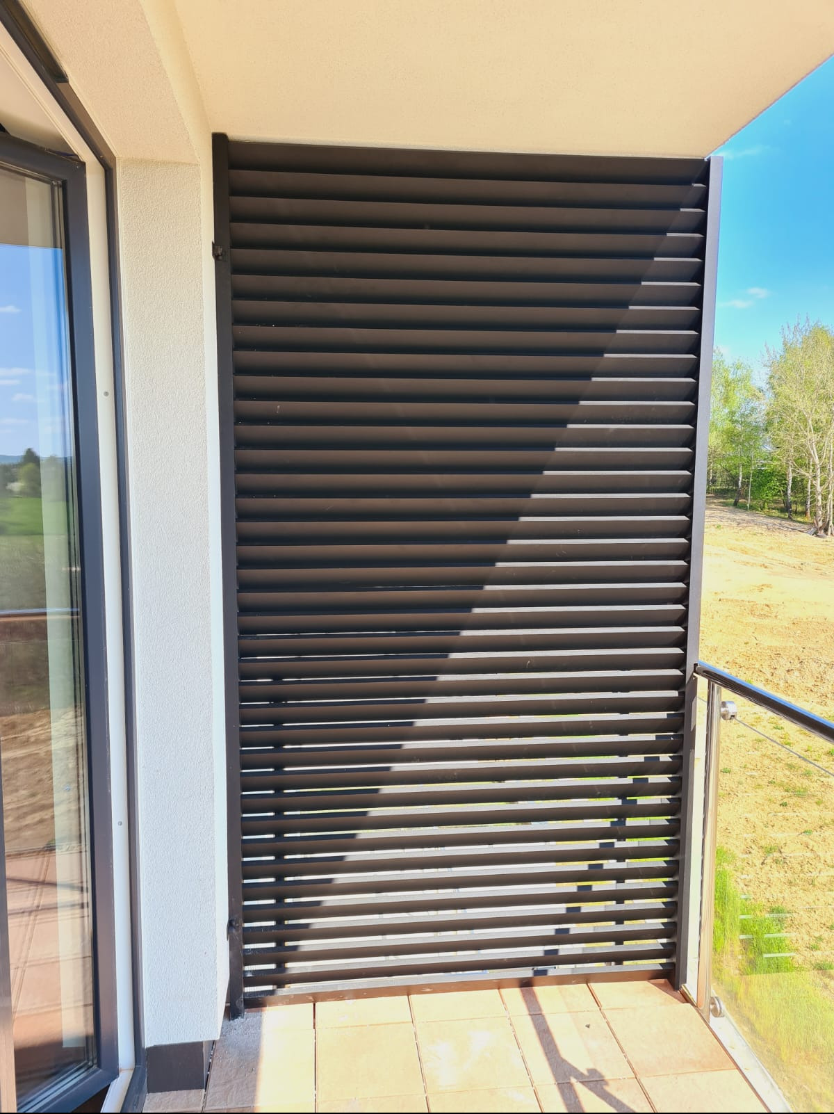 Balkon z nowoczesną, poziomą, metalową przesłoną w kolorze antracytowym, zapewniającą prywatność, z widokiem na zielone wzgórza i błękitne niebo.