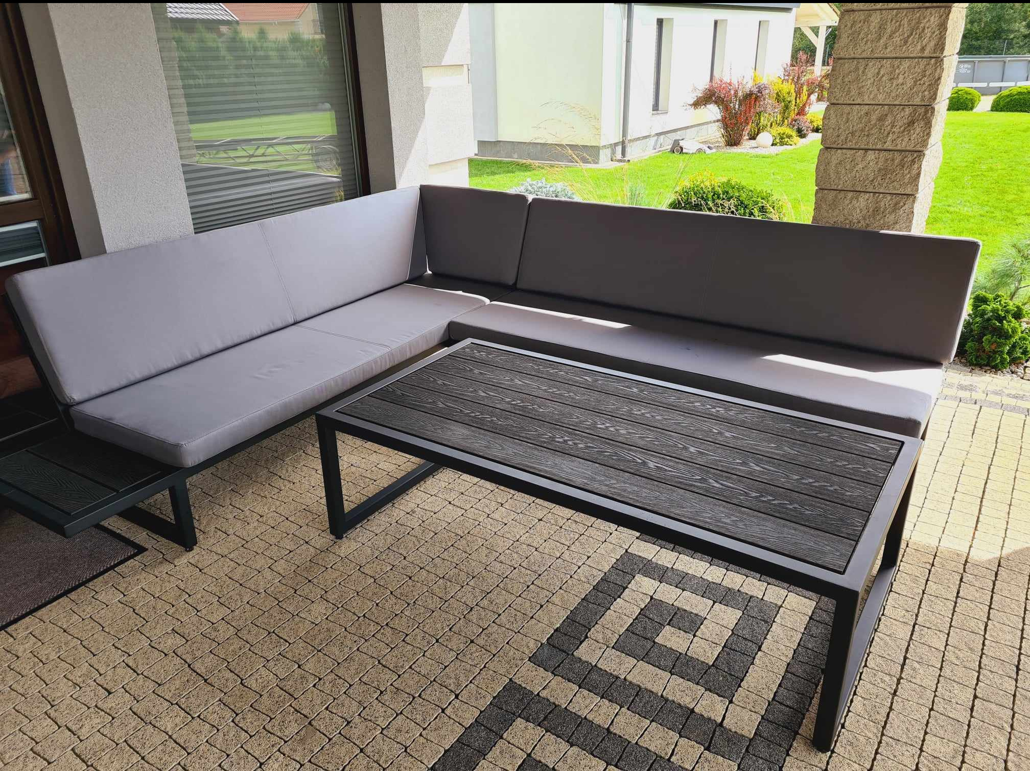 Zadaszony taras z narożną sofą z szarymi poduszkami i stolikiem kawowym z blatem imitującym drewno, na kostce brukowej z geometrycznym wzorem.