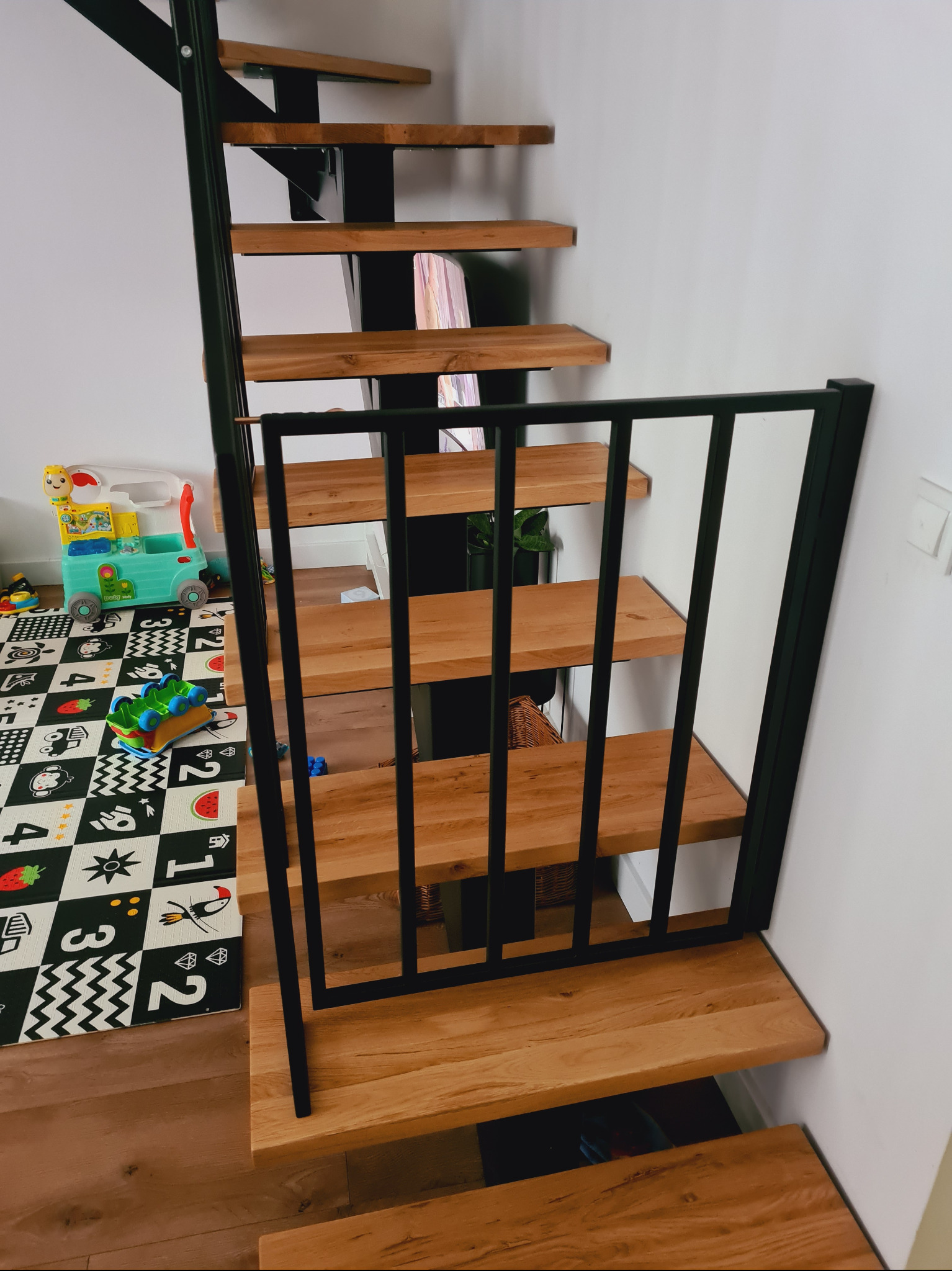 Drewniane schody z czarną metalową konstrukcją i zabezpieczeniem dla dzieci, widoczne zabawki na macie edukacyjnej w tle.