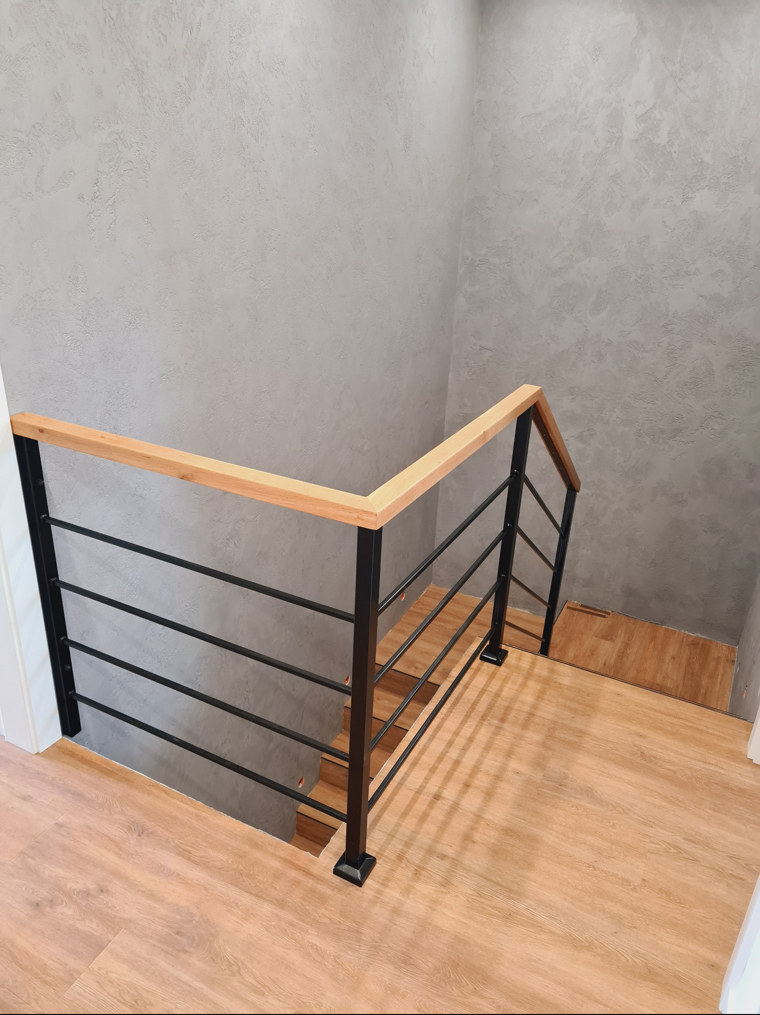 Minimalistyczna balustrada schodowa z czarnymi metalowymi słupkami i poziomymi prętami oraz drewnianym poręczą, widok z góry na drewniane schody i szarą ścianę o fakturze.