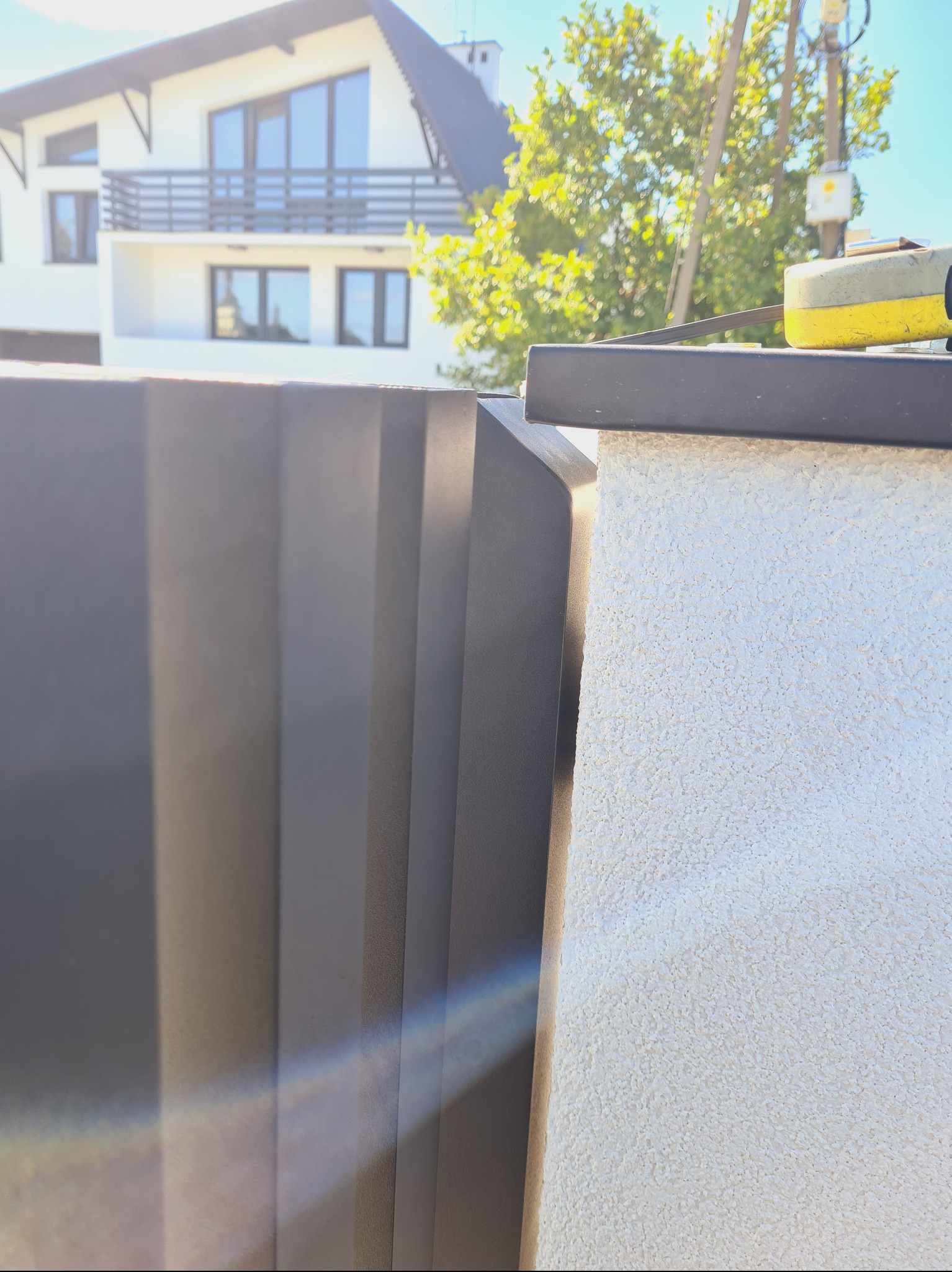 Fragment nowoczesnego ogrodzenia z pionowych, matowych paneli w grafitowym kolorze, przylegającego do ściany z jasnym, ziarnistym tynkiem. Na górze ogrodzenia widoczna żółta miara zwijana. W tle...