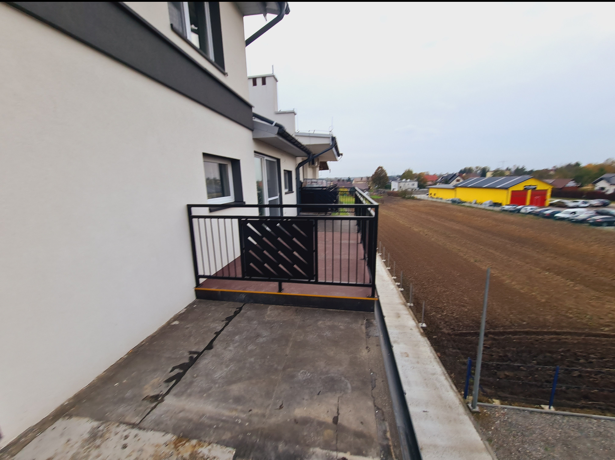 Czarna balustrada balkonowa z dekoracyjnym wypełnieniem w postaci ukośnych prętów, widok z boku na balkon, w tle pole i budynek gospodarczy.