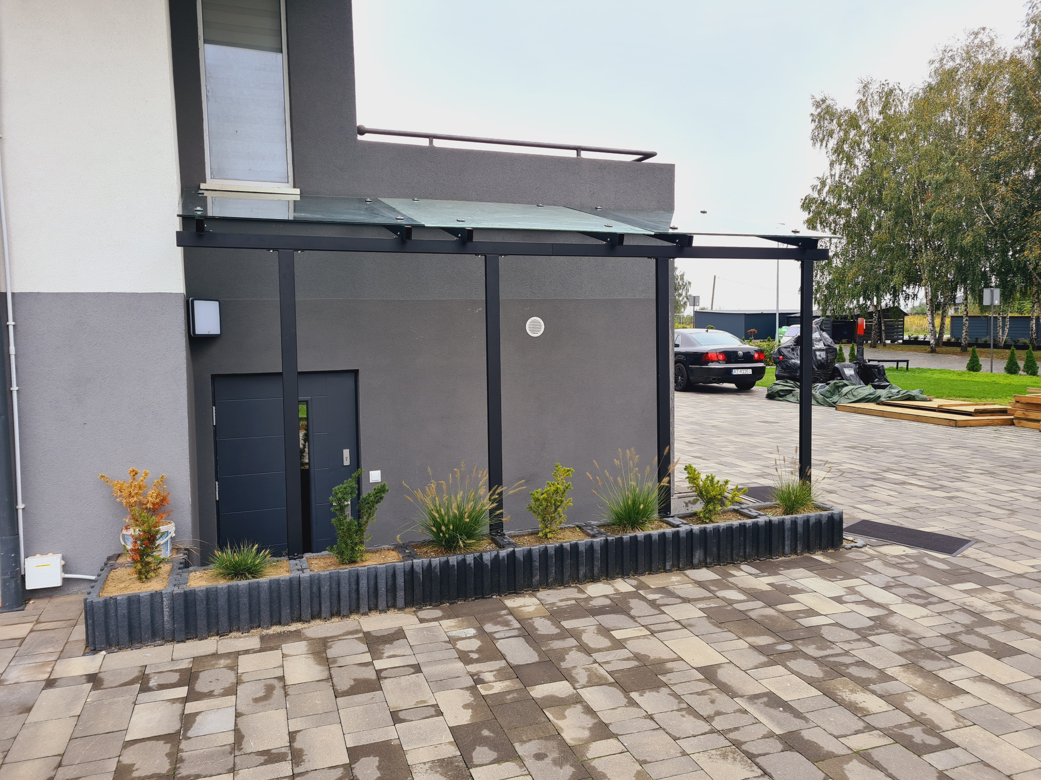 Nowoczesna pergola z czarną metalową konstrukcją i szklanym zadaszeniem przy szarym budynku, otoczona kostką brukową i niskimi rabatami z roślinami ozdobnymi.