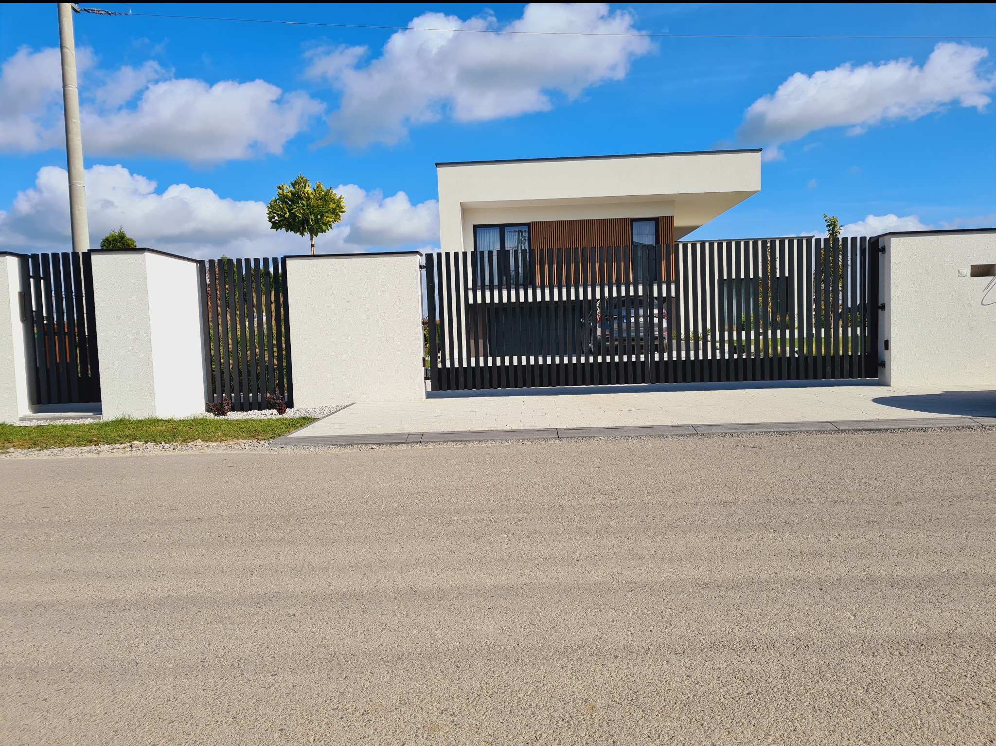 Nowoczesne ogrodzenie posesji z przesuwną bramą, pionowe czarne sztachety, białe murowane słupki, dom w tle, błękitne niebo z chmurami.