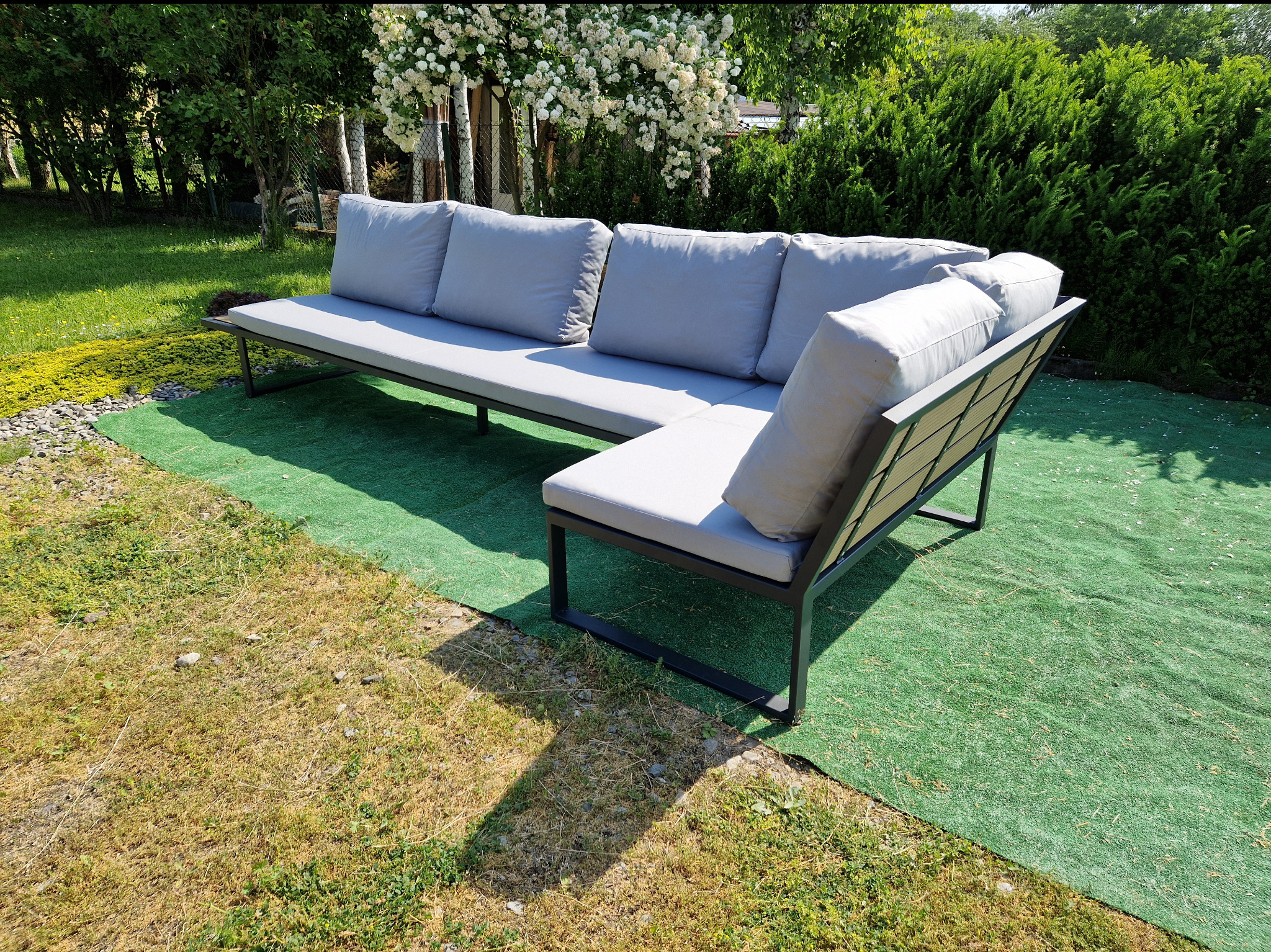 Nowoczesna, narożna sofa ogrodowa z metalową ramą i szarymi poduszkami na tle zieleni ogrodowej i kwitnącego krzewu.