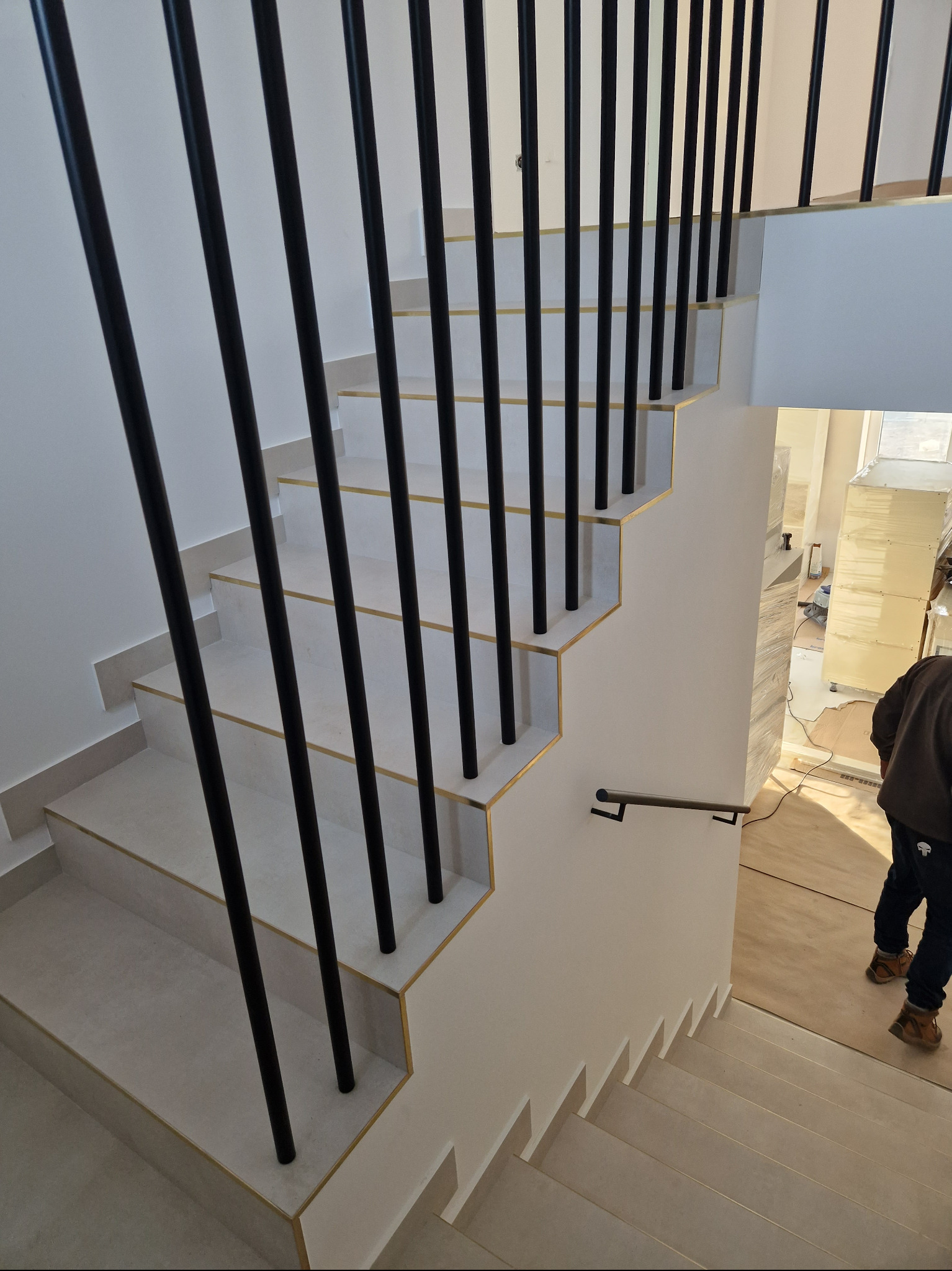 Nowoczesne schody z jasnymi stopniami, czarnymi, pionowymi słupkami balustrady i złotymi akcentami na krawędziach stopni, widok z góry na dół.