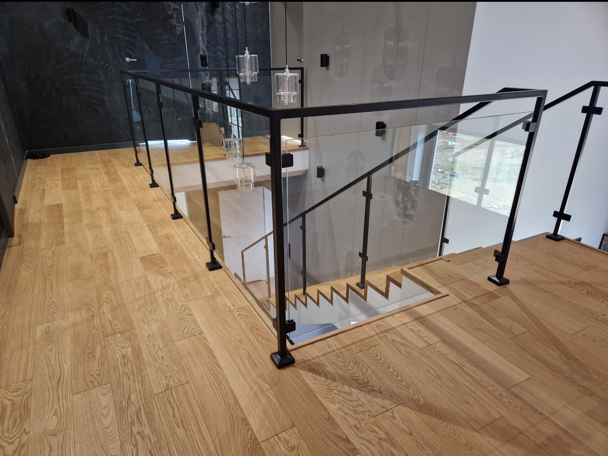Nowoczesna balustrada ze szkła i czarnych metalowych słupków, zamontowana na drewnianych schodach w jasnym wnętrzu z elementami dekoracyjnymi na ścianach.