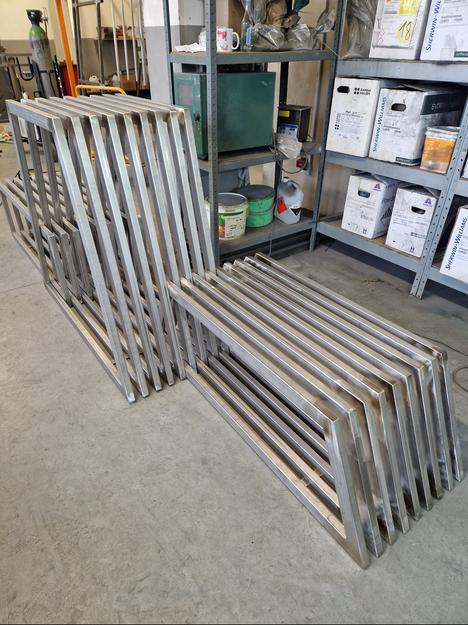 Stos metalowych, spawanych ram o prostokątnym przekroju, prawdopodobnie elementów balustrady lub ogrodzenia, leżących na betonowej podłodze warsztatu w pobliżu regałów z farbami i narzędziami.